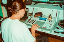 A person with long hair tied back operates music production equipment, including an AKAI MPC and a laptop, in a studio setting. Additional equipment like a turntable and speakers are visible, with various items such as a jar and pens placed on a desk.