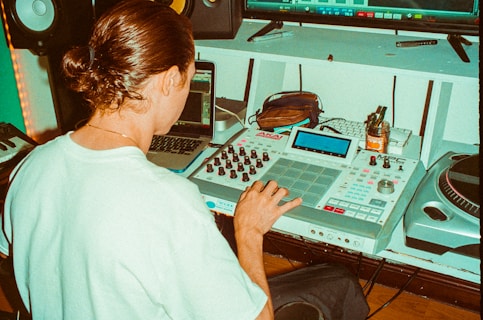 A person with long hair tied back operates music production equipment, including an AKAI MPC and a laptop, in a studio setting. Additional equipment like a turntable and speakers are visible, with various items such as a jar and pens placed on a desk.