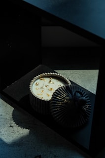 A decorative candle with a round, ribbed black container is topped with a matching lid. The candle is white with gold-colored flecks on the surface, positioned on a dark, shadowy surface.