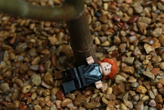 A LEGO figure with red hair and a black suit is lying on a bed of small brown pebbles next to a tree trunk.