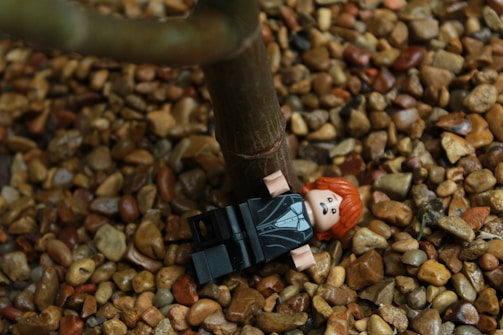 A LEGO figure with red hair and a black suit is lying on a bed of small brown pebbles next to a tree trunk.