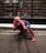 A person in a Spider-Man costume crouches on a sidewalk in front of a storefront displaying multiple shoe boxes. The window has the text 'Converse All Star 100th Anniversary' printed on it. The costume is red and blue with a web pattern, and the pose mimics Spider-Man's signature web-shooting action.