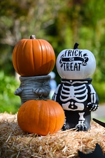 Three pumpkins are arranged on a bed of hay. One is uncarved and placed at the bottom, another is stacked on a figure resembling a mummy with an orange pumpkin on its head, and the third is a white pumpkin with 'TRICK OR TREAT' written on it, placed on a figure with a skeleton body. The background is a garden with green foliage.