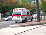 An ambulance is driving at speed through a city street. The vehicle is white with red and blue markings, and features emergency symbols and contact information. Buildings and trees are visible in the background as the scene is slightly blurred, indicating motion.