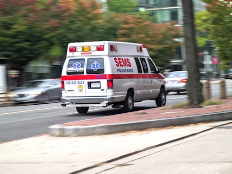 An ambulance is driving at speed through a city street. The vehicle is white with red and blue markings, and features emergency symbols and contact information. Buildings and trees are visible in the background as the scene is slightly blurred, indicating motion.