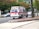 An ambulance is driving at speed through a city street. The vehicle is white with red and blue markings, and features emergency symbols and contact information. Buildings and trees are visible in the background as the scene is slightly blurred, indicating motion.