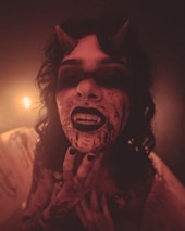A person with horns, wearing dark makeup, appears in a dimly lit environment. Their face and mouth are smeared with a substance resembling blood. The lighting casts a warm glow, adding to the eerie atmosphere.