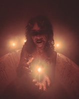 A figure with a sinister and eerie appearance, adorned with small horns, dark makeup, and holding a lit candle. The scene is shrouded in a misty and dim ambiance, intensified by the glow of multiple candles. The background reveals a bathtub with streaks resembling dried blood, enhancing the unsettling atmosphere.