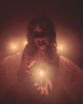 A figure with a sinister and eerie appearance, adorned with small horns, dark makeup, and holding a lit candle. The scene is shrouded in a misty and dim ambiance, intensified by the glow of multiple candles. The background reveals a bathtub with streaks resembling dried blood, enhancing the unsettling atmosphere.