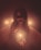 A figure with a sinister and eerie appearance, adorned with small horns, dark makeup, and holding a lit candle. The scene is shrouded in a misty and dim ambiance, intensified by the glow of multiple candles. The background reveals a bathtub with streaks resembling dried blood, enhancing the unsettling atmosphere.