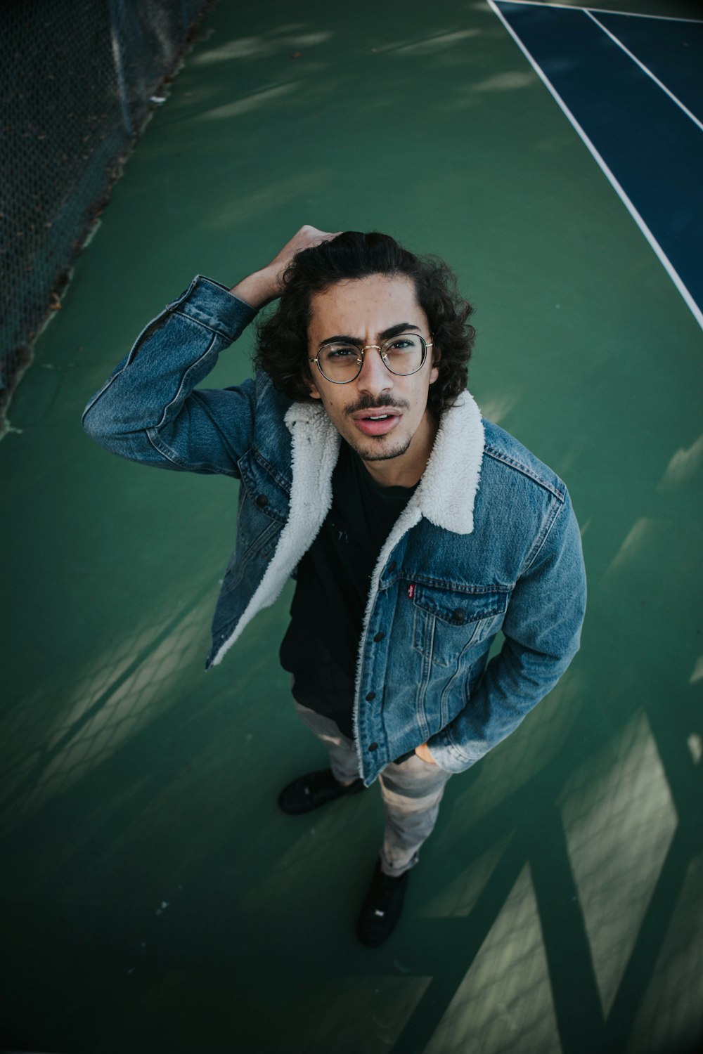 a man with glasses and a denim jacket standing on a tennis court