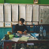 A person is seated at a cluttered desk with papers, bottles, and office supplies. In the background, there is a wall with charts, graphs, and a map. The setting appears to be an office or a planning room.