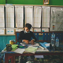 A desk covered with maps, military reports, and a laptop displaying political analysis.