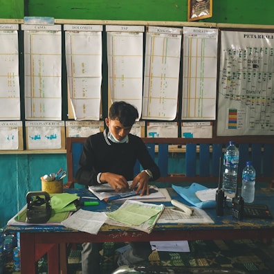 A person is seated at a cluttered desk with papers, bottles, and office supplies. In the background, there is a wall with charts, graphs, and a map. The setting appears to be an office or a planning room.