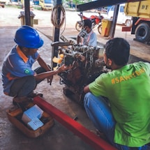Three individuals are working on a mechanical engine under a sheltered area. One is wearing a blue hard hat and examining the engine closely. There are tools and boxes on the ground nearby, and a red metal bar is attached to the engine for support. Various vehicles including a truck and a motorcycle can be seen in the background.