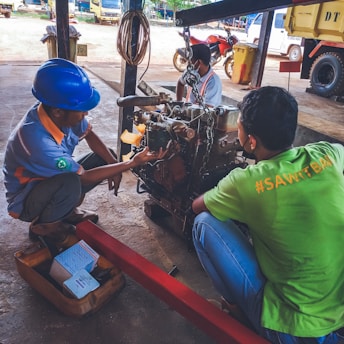 Three individuals are working on a mechanical engine under a sheltered area. One is wearing a blue hard hat and examining the engine closely. There are tools and boxes on the ground nearby, and a red metal bar is attached to the engine for support. Various vehicles including a truck and a motorcycle can be seen in the background.