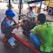 Three individuals are working on a mechanical engine under a sheltered area. One is wearing a blue hard hat and examining the engine closely. There are tools and boxes on the ground nearby, and a red metal bar is attached to the engine for support. Various vehicles including a truck and a motorcycle can be seen in the background.