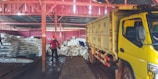 Inside a warehouse, several large stacks of white sacks are piled on the floor. In the foreground, a large yellow truck is parked, partially loaded or unloading. Two people can be seen; one is standing, wearing a red shirt and mask, while the other appears to be organizing or inspecting the bags. The warehouse structure is made of wooden beams painted in red, and the floor appears muddy or wet.