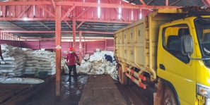 Inside a warehouse, several large stacks of white sacks are piled on the floor. In the foreground, a large yellow truck is parked, partially loaded or unloading. Two people can be seen; one is standing, wearing a red shirt and mask, while the other appears to be organizing or inspecting the bags. The warehouse structure is made of wooden beams painted in red, and the floor appears muddy or wet.