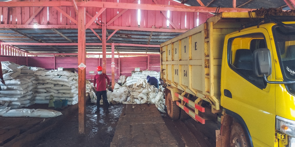 Inside a warehouse, several large stacks of white sacks are piled on the floor. In the foreground, a large yellow truck is parked, partially loaded or unloading. Two people can be seen; one is standing, wearing a red shirt and mask, while the other appears to be organizing or inspecting the bags. The warehouse structure is made of wooden beams painted in red, and the floor appears muddy or wet.