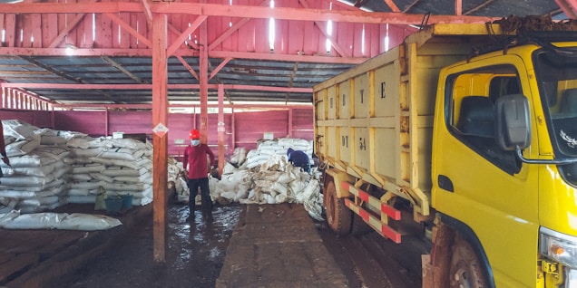 Inside a warehouse, several large stacks of white sacks are piled on the floor. In the foreground, a large yellow truck is parked, partially loaded or unloading. Two people can be seen; one is standing, wearing a red shirt and mask, while the other appears to be organizing or inspecting the bags. The warehouse structure is made of wooden beams painted in red, and the floor appears muddy or wet.