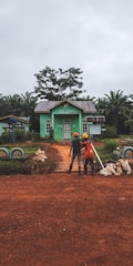 Two individuals wearing construction helmets stand on a dirt path leading to a small, green building surrounded by lush greenery and palm trees. The building has a metal roof and a sign above the door. The scene conveys a rural or agricultural setting, with construction materials and dirt bags visible around the path.
