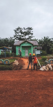 Two individuals wearing construction helmets stand on a dirt path leading to a small, green building surrounded by lush greenery and palm trees. The building has a metal roof and a sign above the door. The scene conveys a rural or agricultural setting, with construction materials and dirt bags visible around the path.