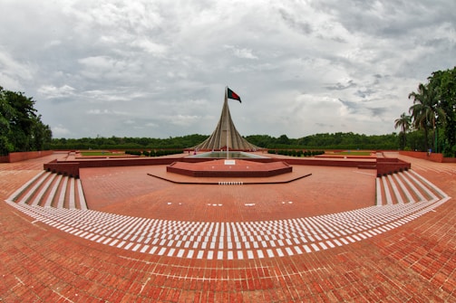 A striking monument stands in a large open area, featuring a tall, triangular structure with a flag flying at the top. The ground is paved with red bricks, and symmetrical, wide staircases with white markings lead up to the monument. Lush green trees surround the area under a cloudy sky.