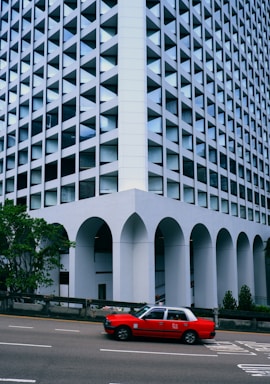 A red taxi drives past a geometric building with a unique facade featuring repeating triangular patterns and large arches at the base. The building has a modern, architectural design with reflective glass, and it is flanked by some greenery.