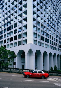 A red taxi drives past a geometric building with a unique facade featuring repeating triangular patterns and large arches at the base. The building has a modern, architectural design with reflective glass, and it is flanked by some greenery.