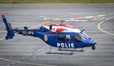 A blue and white police helicopter is parked on a runway with a circular marking visible on the asphalt. The helicopter displays the word 'POLIS' on the side, indicating it is used for law enforcement, and has the registration number SE-JPV. The aircraft has a number of visible components, including a large rotor blade, doors, and windows.
