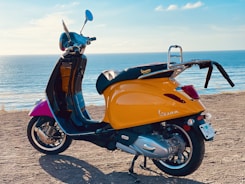 A vibrant yellow Vespa scooter is parked on a sandy path near an ocean. The background consists of a clear blue sky and calm sea waves, creating a serene coastal atmosphere. The scooter is angled to show its side profile, with a rear luggage rack and chrome mirrors adding to its classic design.