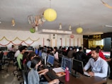 A lively office environment filled with people working at computers. There are festive decorations hanging from the ceiling, including paper lanterns and streamers. The employees are sitting in rows, focused on their screens, with some conducting work on laptops. Bright decorations adorn the walls, creating a vibrant and celebratory atmosphere.