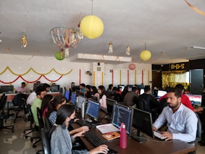 A lively office environment filled with people working at computers. There are festive decorations hanging from the ceiling, including paper lanterns and streamers. The employees are sitting in rows, focused on their screens, with some conducting work on laptops. Bright decorations adorn the walls, creating a vibrant and celebratory atmosphere.