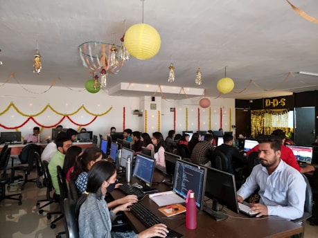 A lively office environment filled with people working at computers. There are festive decorations hanging from the ceiling, including paper lanterns and streamers. The employees are sitting in rows, focused on their screens, with some conducting work on laptops. Bright decorations adorn the walls, creating a vibrant and celebratory atmosphere.