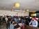 A lively office environment filled with people working at computers. There are festive decorations hanging from the ceiling, including paper lanterns and streamers. The employees are sitting in rows, focused on their screens, with some conducting work on laptops. Bright decorations adorn the walls, creating a vibrant and celebratory atmosphere.