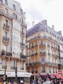 Elegantly designed Parisian buildings with intricate wrought iron balconies line the street. The lower level features cafes and small shops with charming awnings, and people are bustling on the sidewalk.