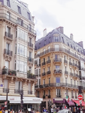 Elegantly designed Parisian buildings with intricate wrought iron balconies line the street. The lower level features cafes and small shops with charming awnings, and people are bustling on the sidewalk.