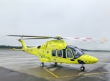 A yellow and green emergency medical helicopter is stationed on a wet asphalt surface, likely an airport or helipad. The helicopter features the word 'AMBULANS' prominently, with additional medical symbols and company markings. The background is overcast, with a cloudy sky and a hint of forest along the horizon.