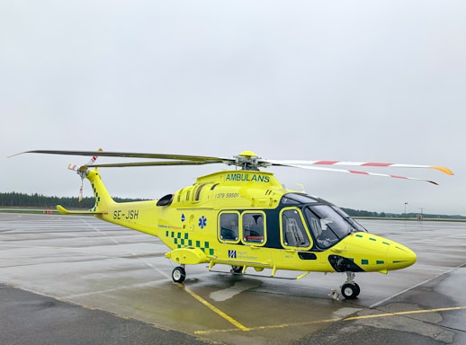 A yellow and green emergency medical helicopter is stationed on a wet asphalt surface, likely an airport or helipad. The helicopter features the word 'AMBULANS' prominently, with additional medical symbols and company markings. The background is overcast, with a cloudy sky and a hint of forest along the horizon.