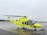 A yellow and green emergency medical helicopter is stationed on a wet asphalt surface, likely an airport or helipad. The helicopter features the word 'AMBULANS' prominently, with additional medical symbols and company markings. The background is overcast, with a cloudy sky and a hint of forest along the horizon.