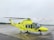 A yellow and green emergency medical helicopter is stationed on a wet asphalt surface, likely an airport or helipad. The helicopter features the word 'AMBULANS' prominently, with additional medical symbols and company markings. The background is overcast, with a cloudy sky and a hint of forest along the horizon.