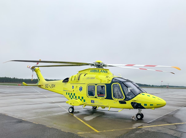 A yellow and green emergency medical helicopter is stationed on a wet asphalt surface, likely an airport or helipad. The helicopter features the word 'AMBULANS' prominently, with additional medical symbols and company markings. The background is overcast, with a cloudy sky and a hint of forest along the horizon.