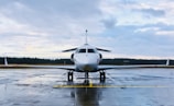 A private jet is parked on a wet runway, reflecting the overcast sky. The aircraft is positioned in the center, with a forest in the distance under a cloudy sky.