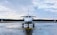 A private jet is parked on a wet runway, reflecting the overcast sky. The aircraft is positioned in the center, with a forest in the distance under a cloudy sky.