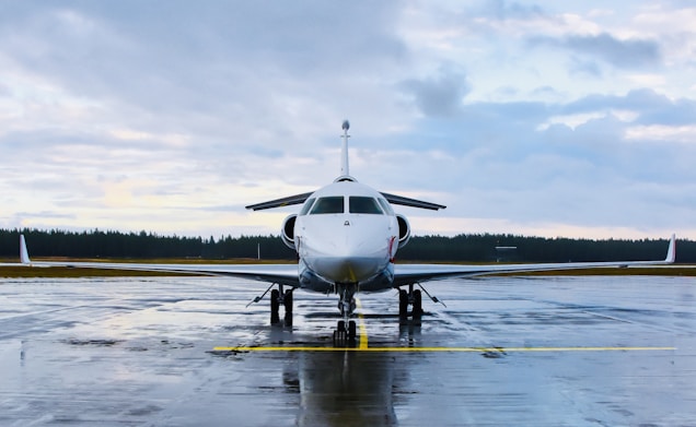 A private jet is parked on a wet runway, reflecting the overcast sky. The aircraft is positioned in the center, with a forest in the distance under a cloudy sky.