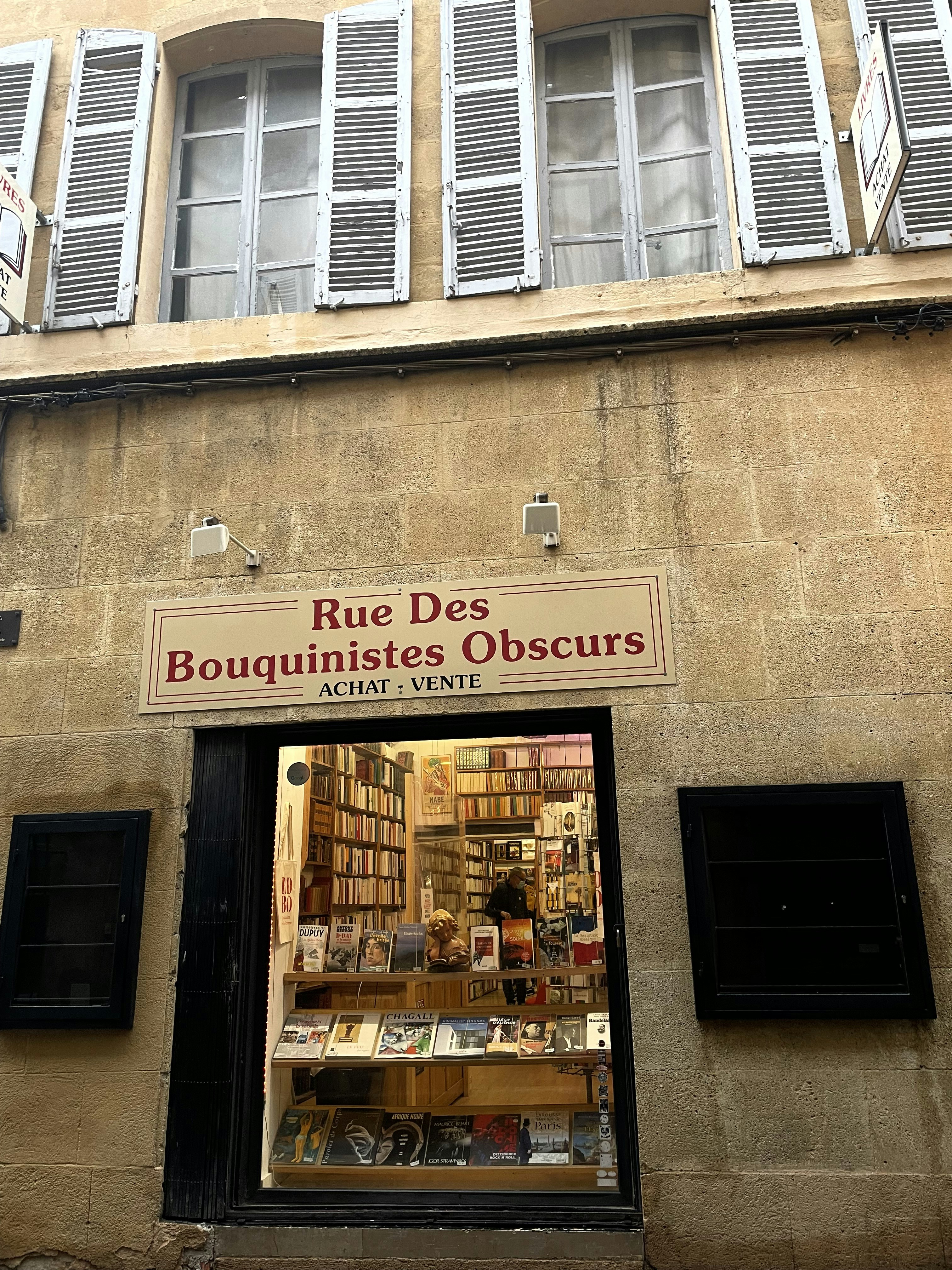 A bookstore display captures the viewer's attention, showcasing a variety of books in the window. The exterior wall features cream-colored stone with traditional white and green shutters framing the windows above. The bookstore sign reads 'Rue Des Bouquinistes Obscurs ACHAT : VENTE'. Through the glass, rows of books line the shelves, with a few prominent titles on display at the front.