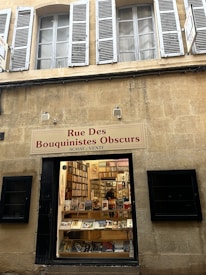 A bookstore display captures the viewer's attention, showcasing a variety of books in the window. The exterior wall features cream-colored stone with traditional white and green shutters framing the windows above. The bookstore sign reads 'Rue Des Bouquinistes Obscurs ACHAT : VENTE'. Through the glass, rows of books line the shelves, with a few prominent titles on display at the front.