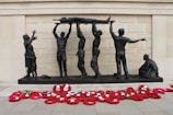 A group of bronze sculptures depicting soldiers hoisting a wounded comrade on a stretcher against the backdrop of a memorial wall. A woman and child stand nearby, while another soldier kneels on the ground. Bright red poppy wreaths are arranged in front of the memorial.