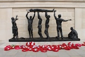 A group of bronze sculptures depicting soldiers hoisting a wounded comrade on a stretcher against the backdrop of a memorial wall. A woman and child stand nearby, while another soldier kneels on the ground. Bright red poppy wreaths are arranged in front of the memorial.